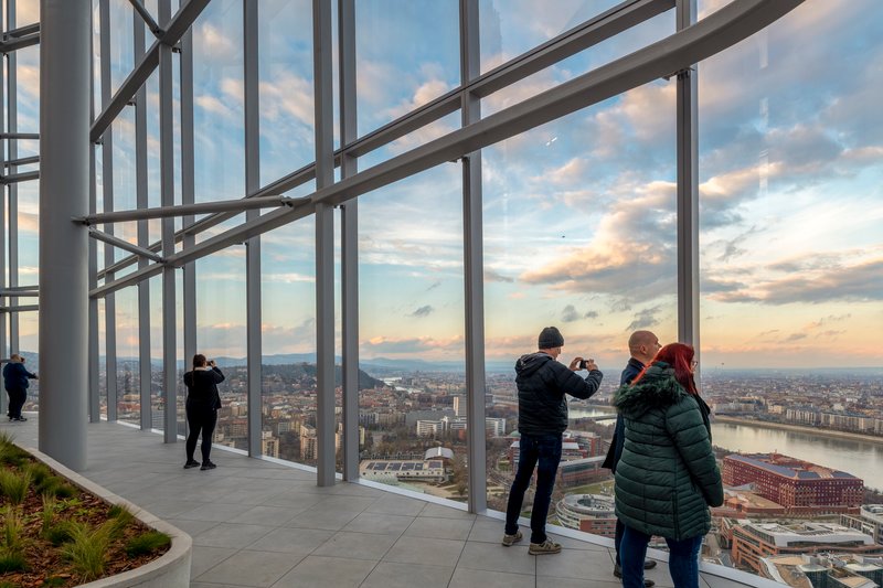 Floor-to-ceiling glazed observation deck with white steel bracing and visitors photographing the cityscape at sunset