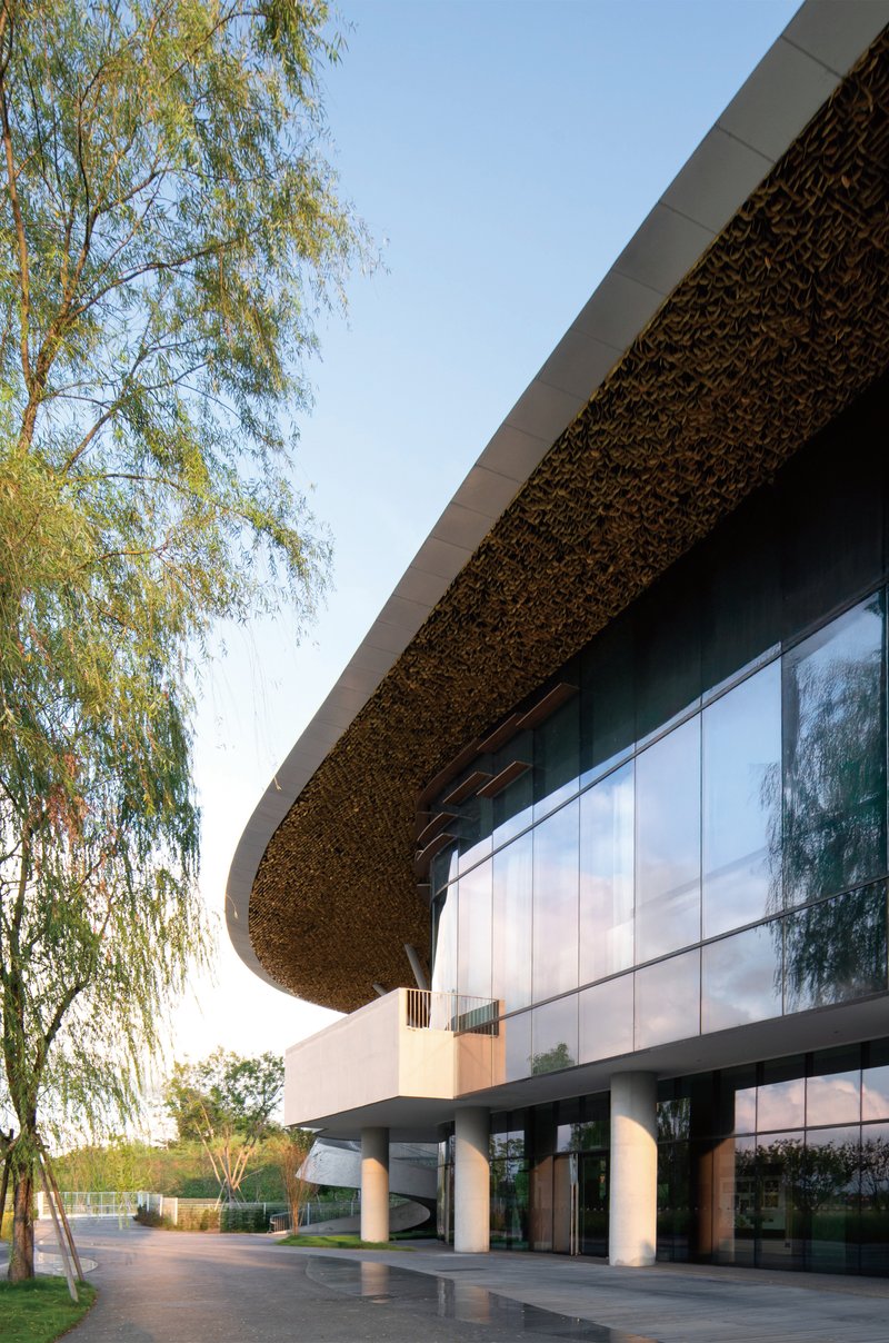 Glazed facade beneath a textured undulating roof canopy with white columns and willow trees at dusk