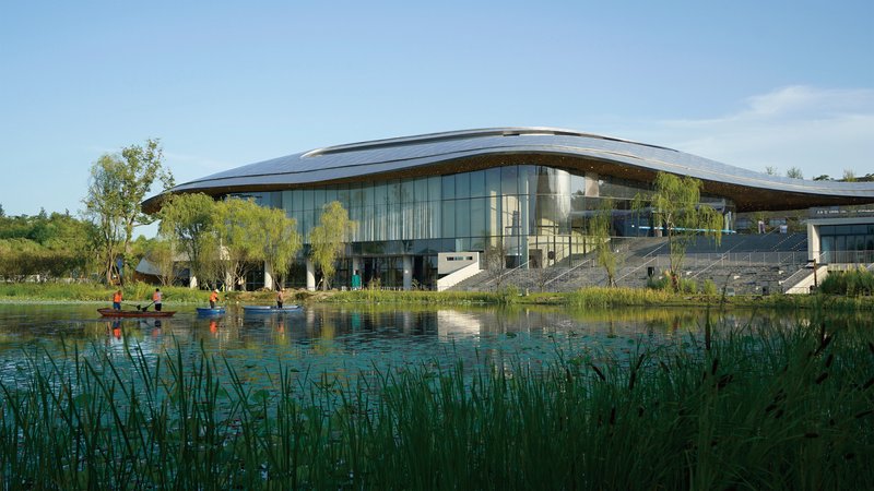 Glass curtain wall and curved roof reflected in a pond edged with tall grasses and visitors