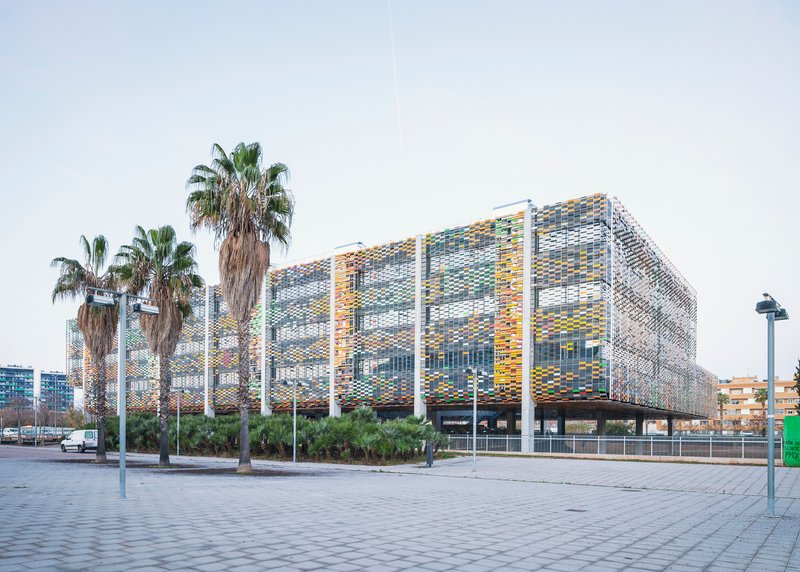 Perforated facade with multicoloured ceramic panels on white columns viewed from a paved plaza with palm trees