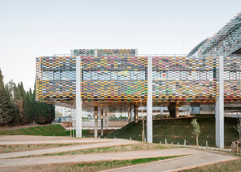 Elevated structure with multicoloured ceramic screen cladding supported by white concrete columns above a landscaped slope
