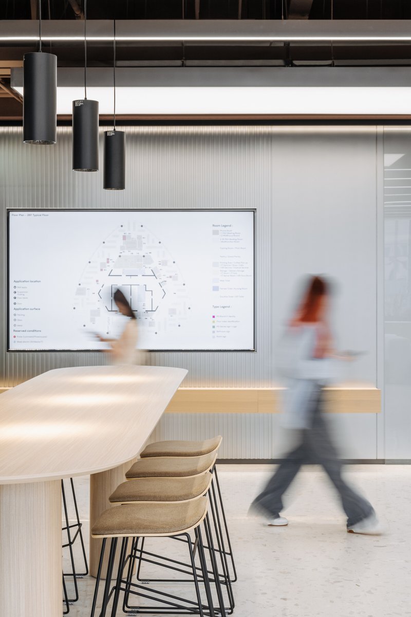 Meeting room with a long table and blurred figures walking past a wall-mounted digital display