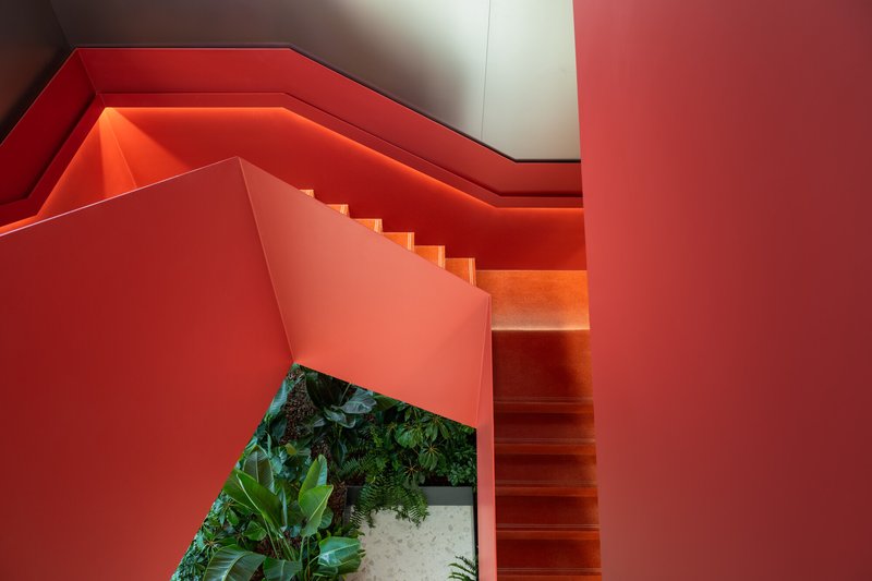 Orange-lit stairwell with folded red walls descending past a ground-level planter bed with ferns and tropical plants