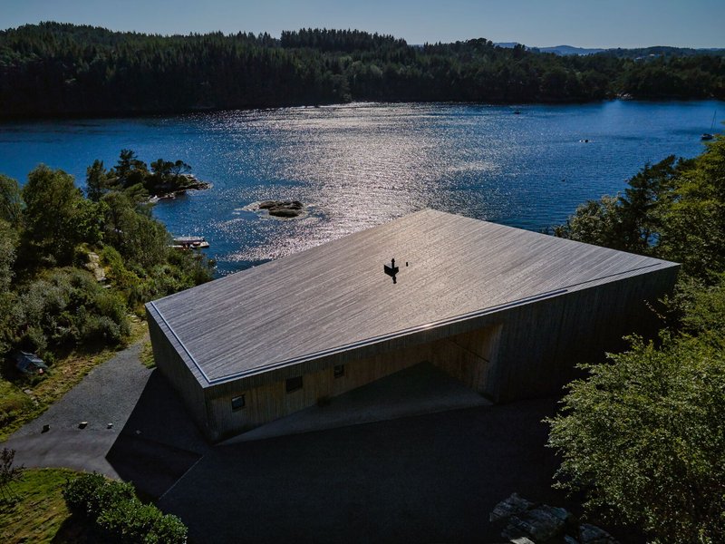 Aerial view of the low-pitched timber roof extending over the structure beside a sunlit fjord