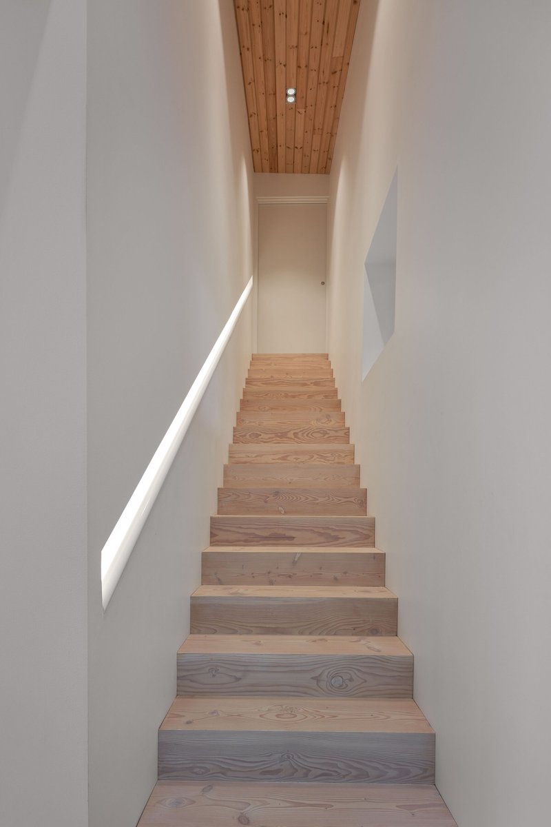 Timber staircase ascending between white walls with a wood-paneled ceiling and slit window