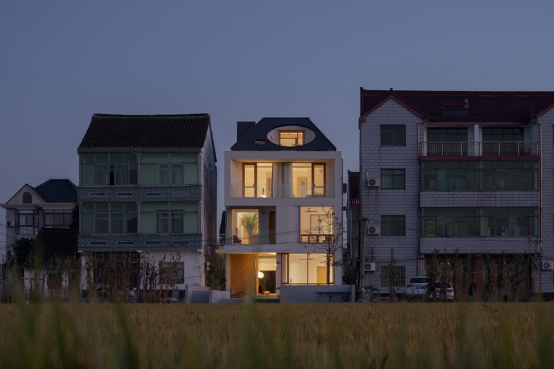 White rendered facade with illuminated arched dormer window nestled among neighboring houses at dusk