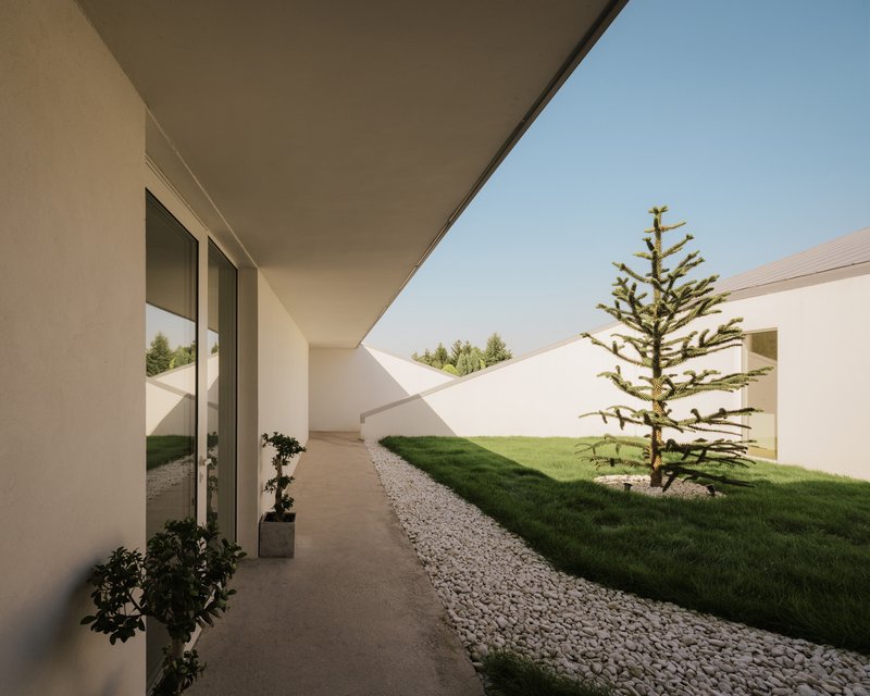 Covered walkway along courtyard edge with conifer tree and gravel strips between grass