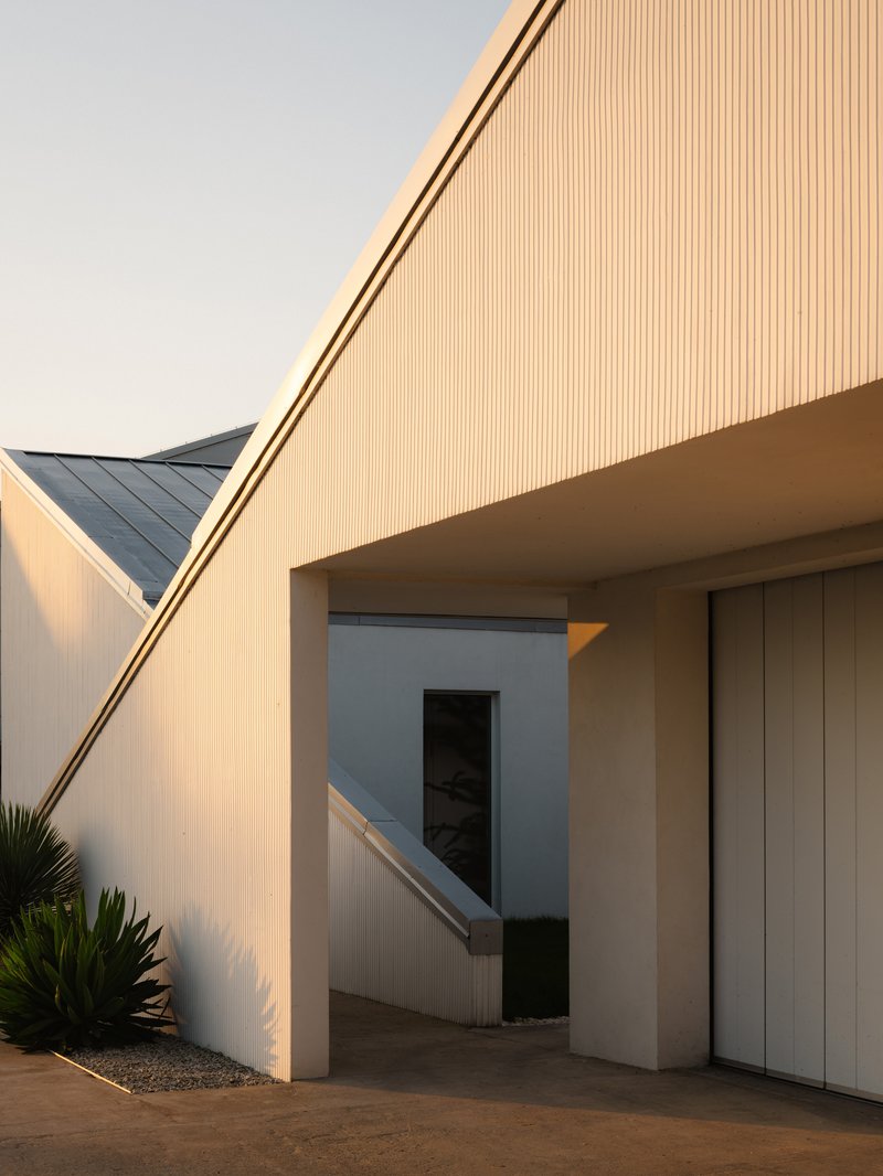 Sloped vertical-ribbed soffit over a courtyard passage at golden hour with planted gravel bed