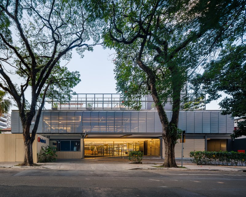 Street facade with translucent upper level, timber cladding at entry, and mature trees flanking the sidewalk at dusk