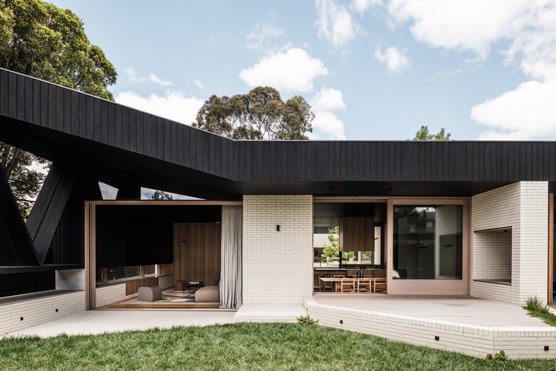 Rear courtyard with white brick walls, timber-framed glazing and a cantilevered black timber roof under midday sun