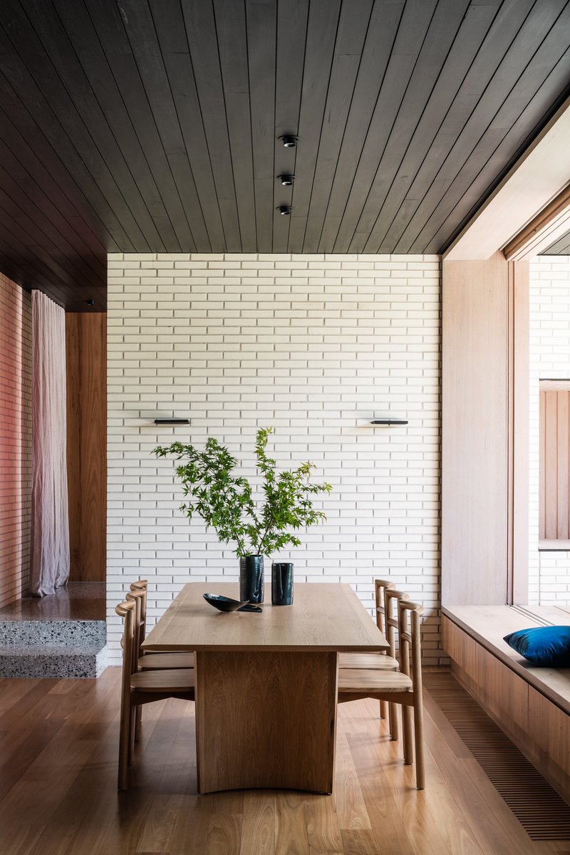 Dining space with timber table and chairs against cream brick wall under black timber ceiling with skylight