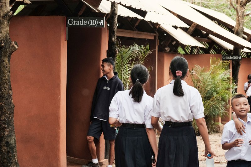 Students gathering outside classroom entries marked with grade numbers under the sloped corrugated roof