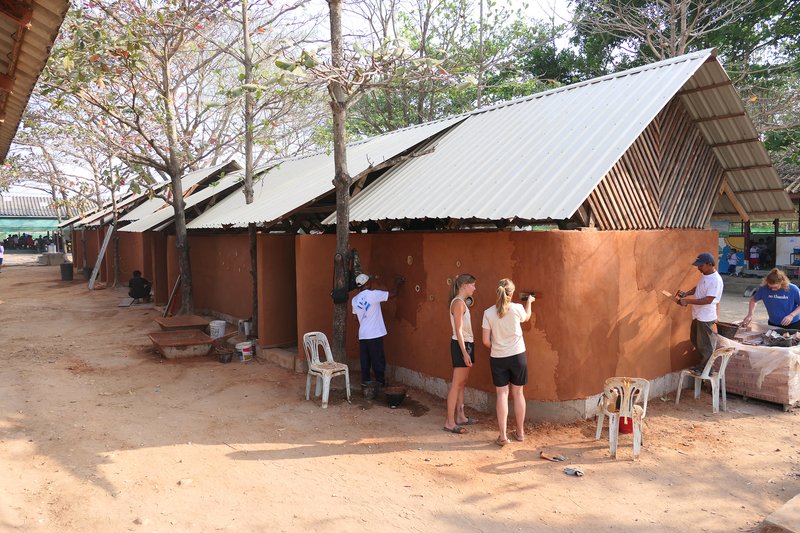 Exterior view of red ochre walls with corrugated metal roofs and visitors gathering in sandy courtyard