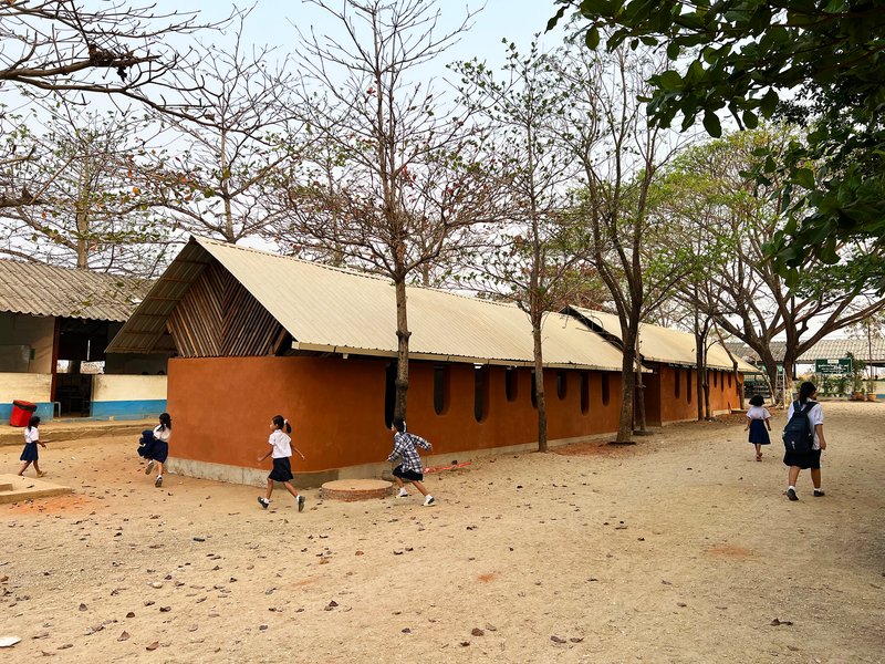 Rammed earth classroom block with corrugated metal gable roof and children running across a sandy courtyard