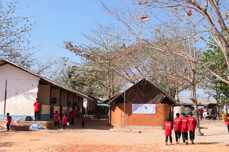 School courtyard showing rammed earth pavilion with pitched roof surrounded by bare deciduous trees and students in red uniforms