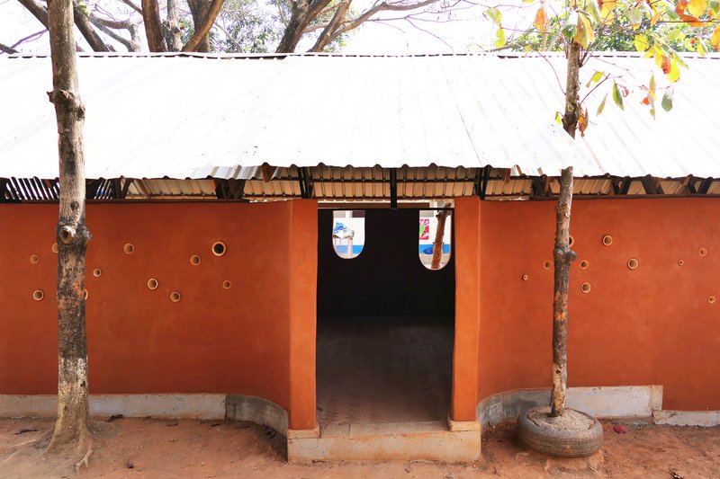 Orange rammed earth wall with circular brass porthole openings flanking a central doorway, framed by two trees