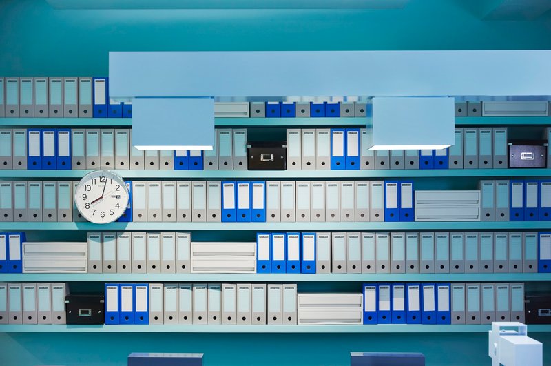 Wall-mounted shelving system displaying rows of blue and grey filing binders with a round clock