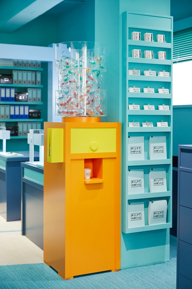 Yellow pedestal dispenser with transparent acrylic top filled with colorful objects beside turquoise shelving unit