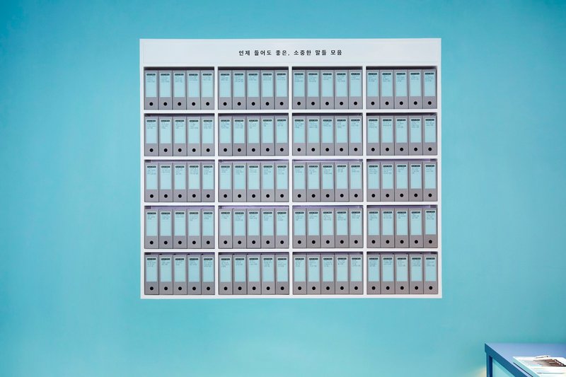 Wall-mounted grid of labeled binder boxes arranged in five rows against a teal surface