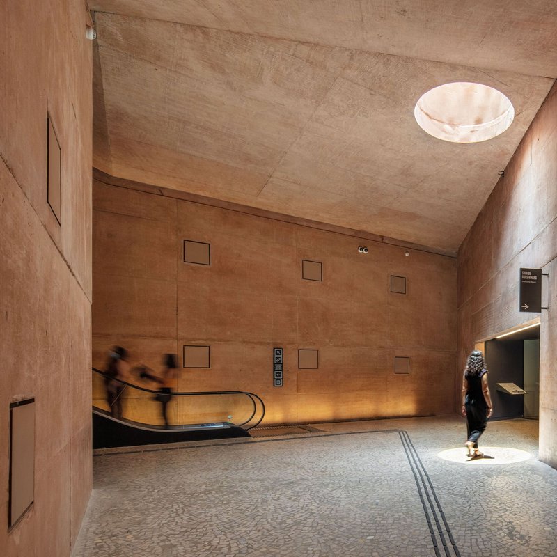 Rammed earth lobby with circular skylight, escalator, and blurred figures in motion across terrazzo floor