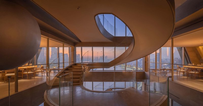 Helical staircase rising through two dining levels with floor-to-ceiling windows overlooking the cityscape at dusk