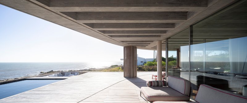 Covered terrace with concrete ceiling beams and perforated timber screens overlooking an infinity pool and coastline