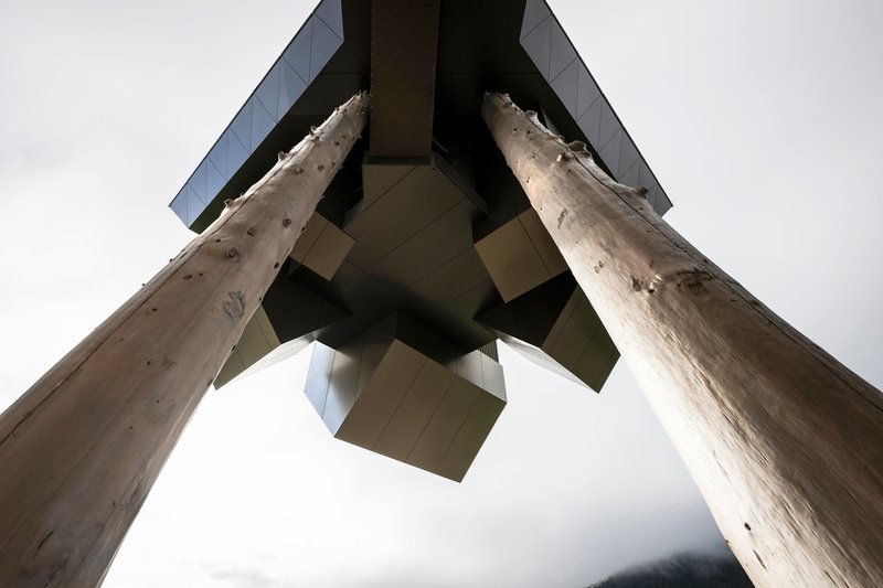 Looking up at the junction of two raw timber columns supporting the angular metal-clad structure