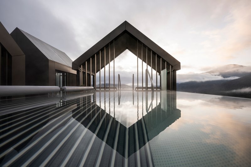 Gabled pavilion with vertical slat screen reflected in still water under overcast mountain sky