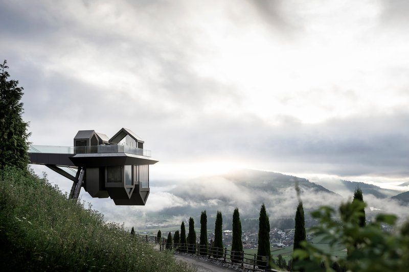 Cantilevered structure with gabled volumes suspended over a misty alpine valley lined with cypress trees