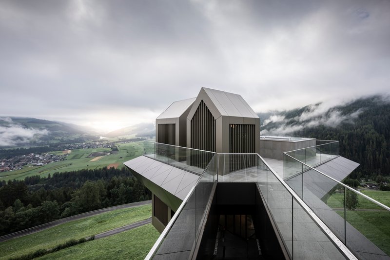 Three dark gabled volumes with vertical cladding atop a faceted metal base under heavy cloud cover