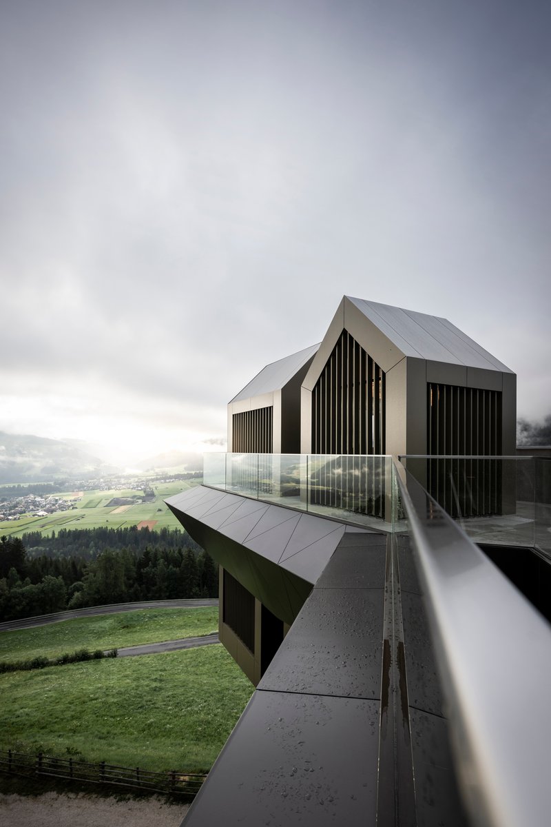 Corner view of the glazed platform with folded metal edges and gabled pavilions above the hillside