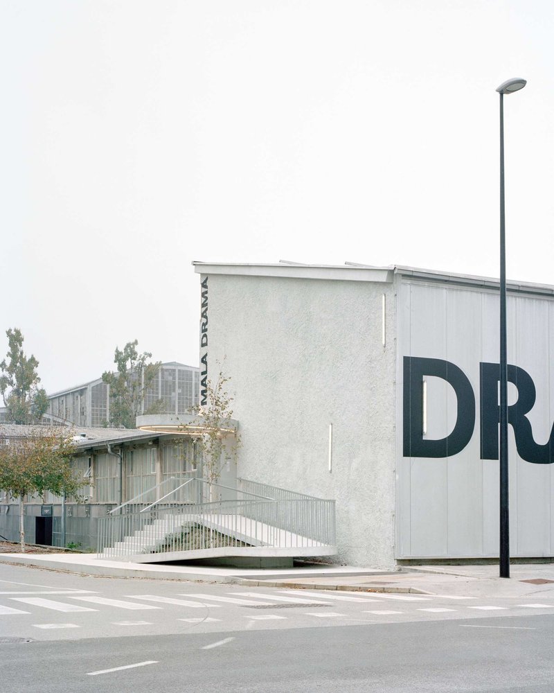 Street view of the stucco volume with large lettering and ramped walkway beside a lamppost