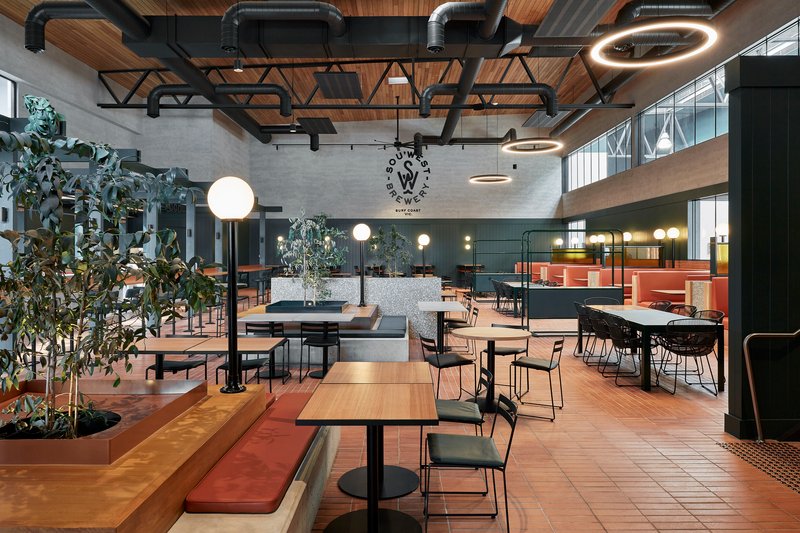 Taproom interior with exposed black steel trusses, timber ceiling panels and potted trees beside terracotta tile floor