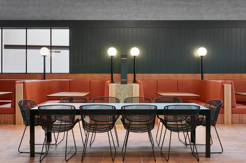 Communal dining table with wire chairs and terrazzo plinth beneath globe pendant lights at dusk
