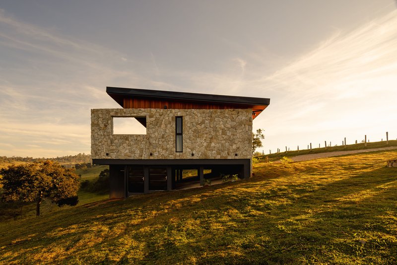 Two-story volume with stacked stone upper level and recessed glazed base on a grassy hillside at dusk