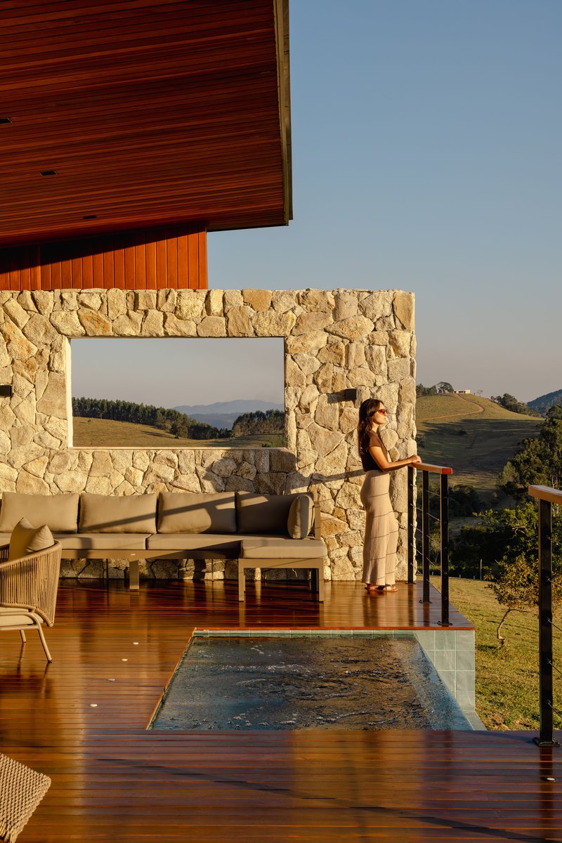 Covered timber deck with stone-framed opening and a person standing by the glass railing at sunset