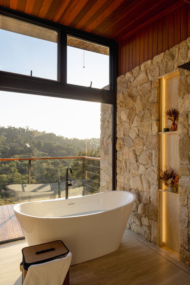 Freestanding bathtub beside a rough stone wall with illuminated niches and views to forested hills