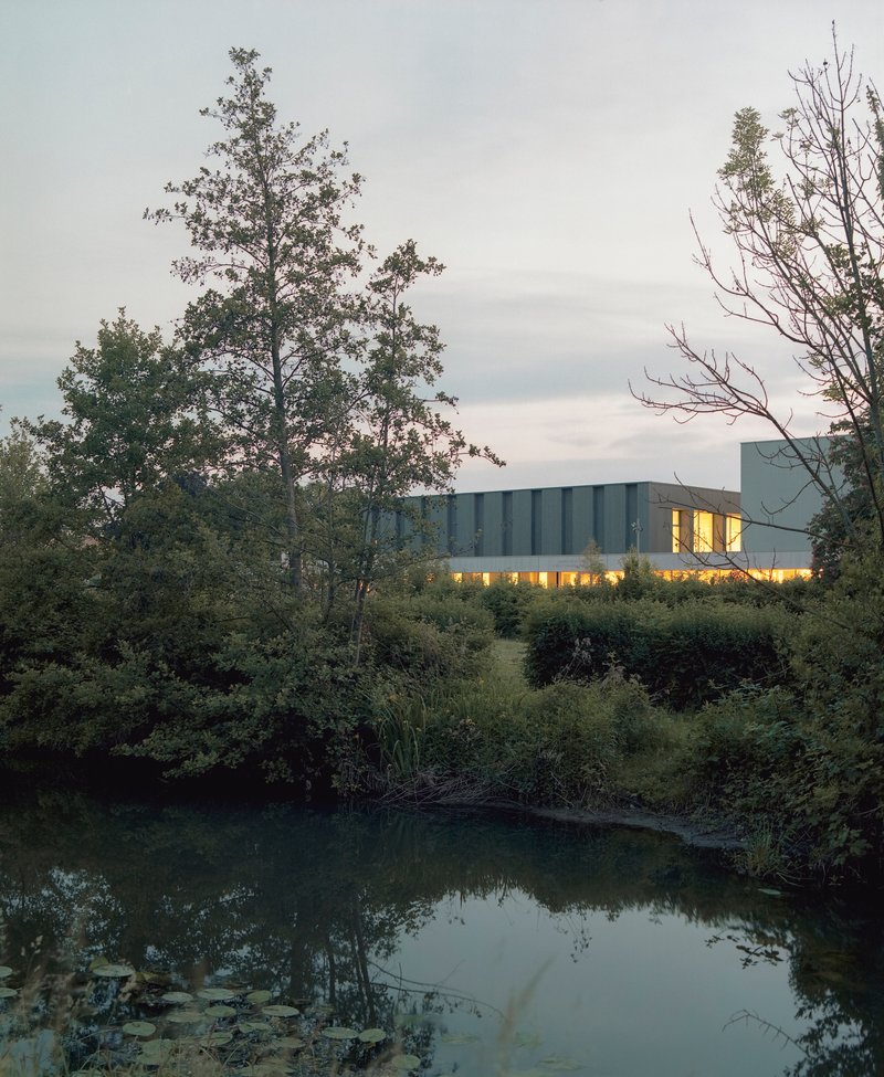 Illuminated upper volume with ribbed facade framed by trees and pond at dusk
