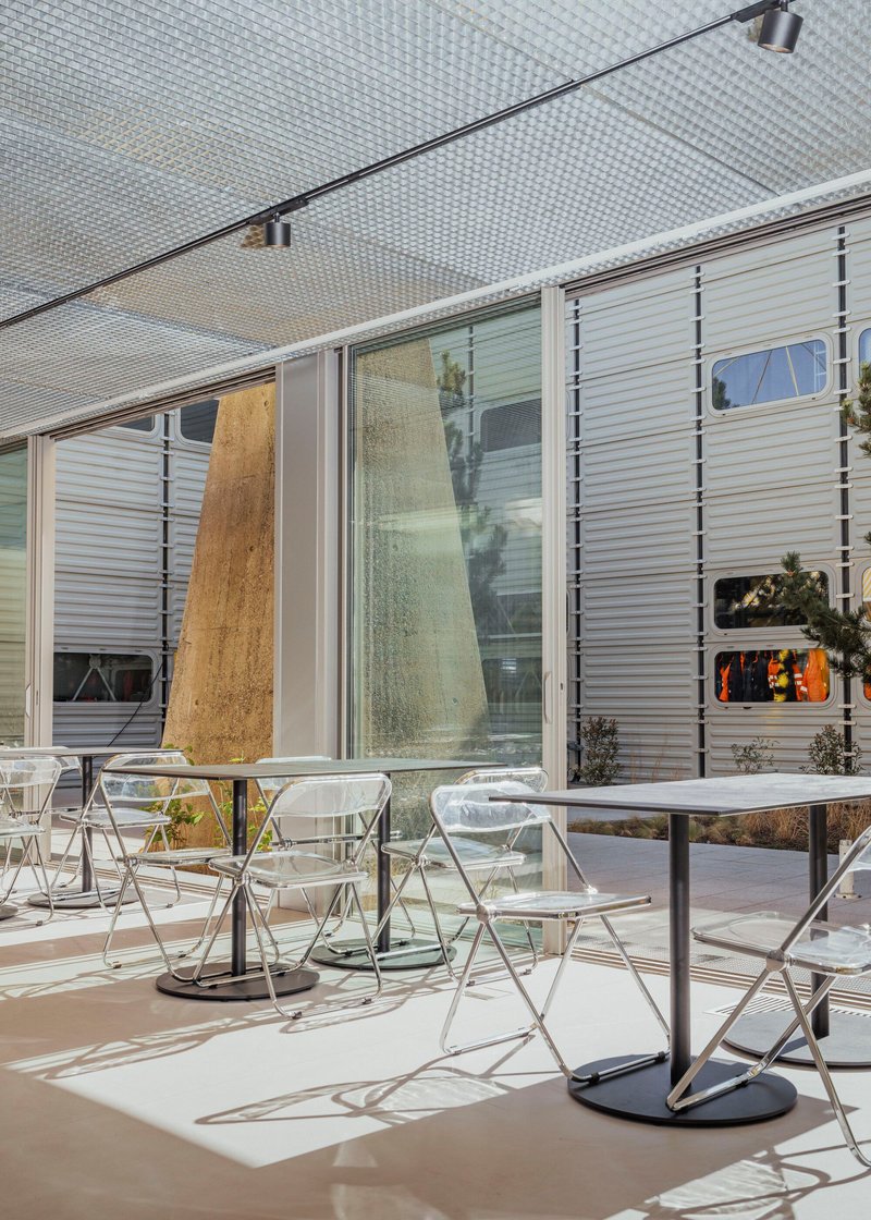 Outdoor seating terrace beneath perforated metal canopy with corrugated facade panels and planted beds in afternoon light