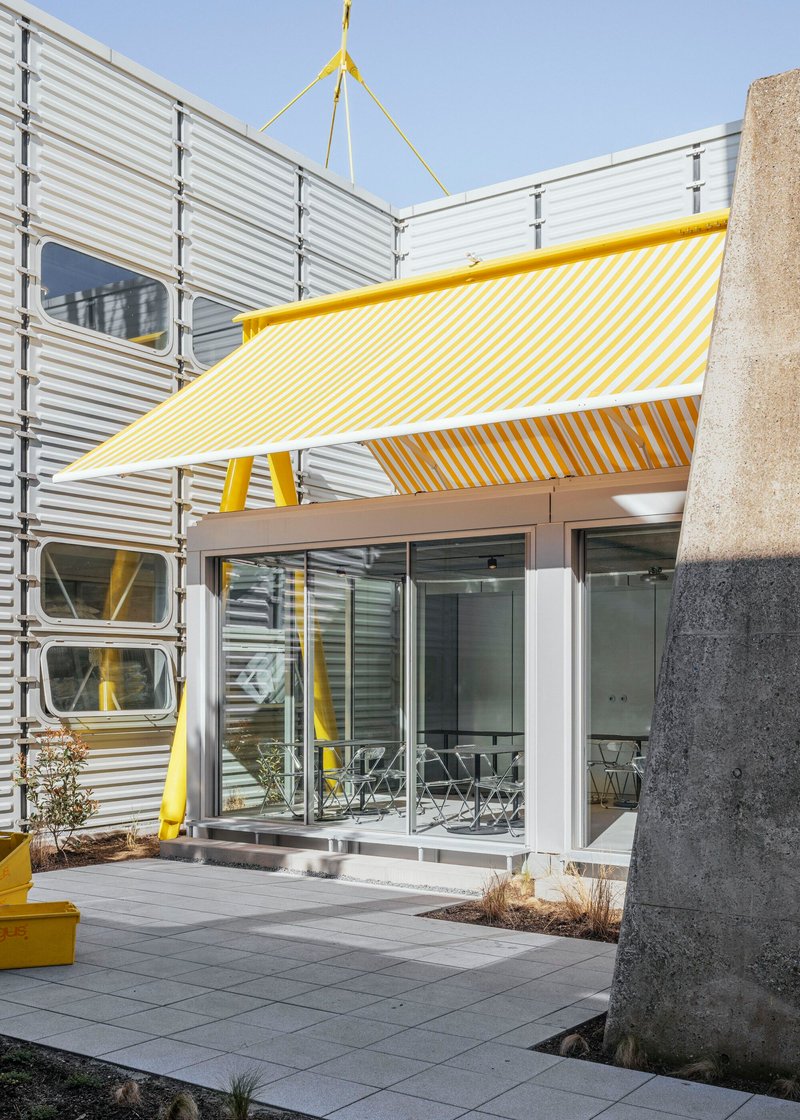 Covered entry threshold with yellow striped fabric canopy, concrete column, and corrugated metal facade under blue sky