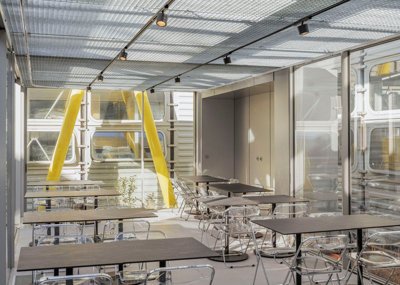 Interior dining area with translucent corrugated ceiling panels, track lighting, and yellow structural columns visible through glazing