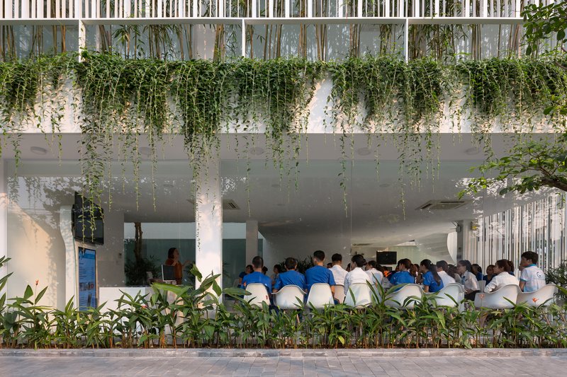 Open-air ground floor with students seated beneath a planted concrete slab and vertical bamboo screens