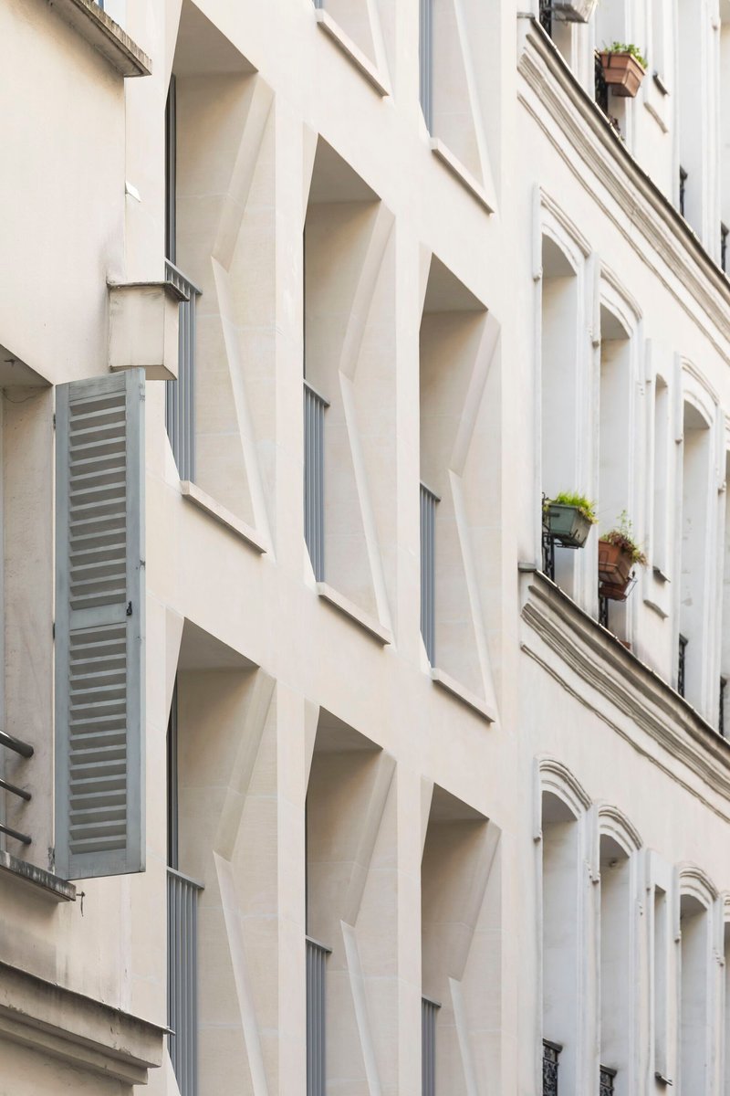 Close-up of white facade showing tapered window openings and grey louvered shutter at corner