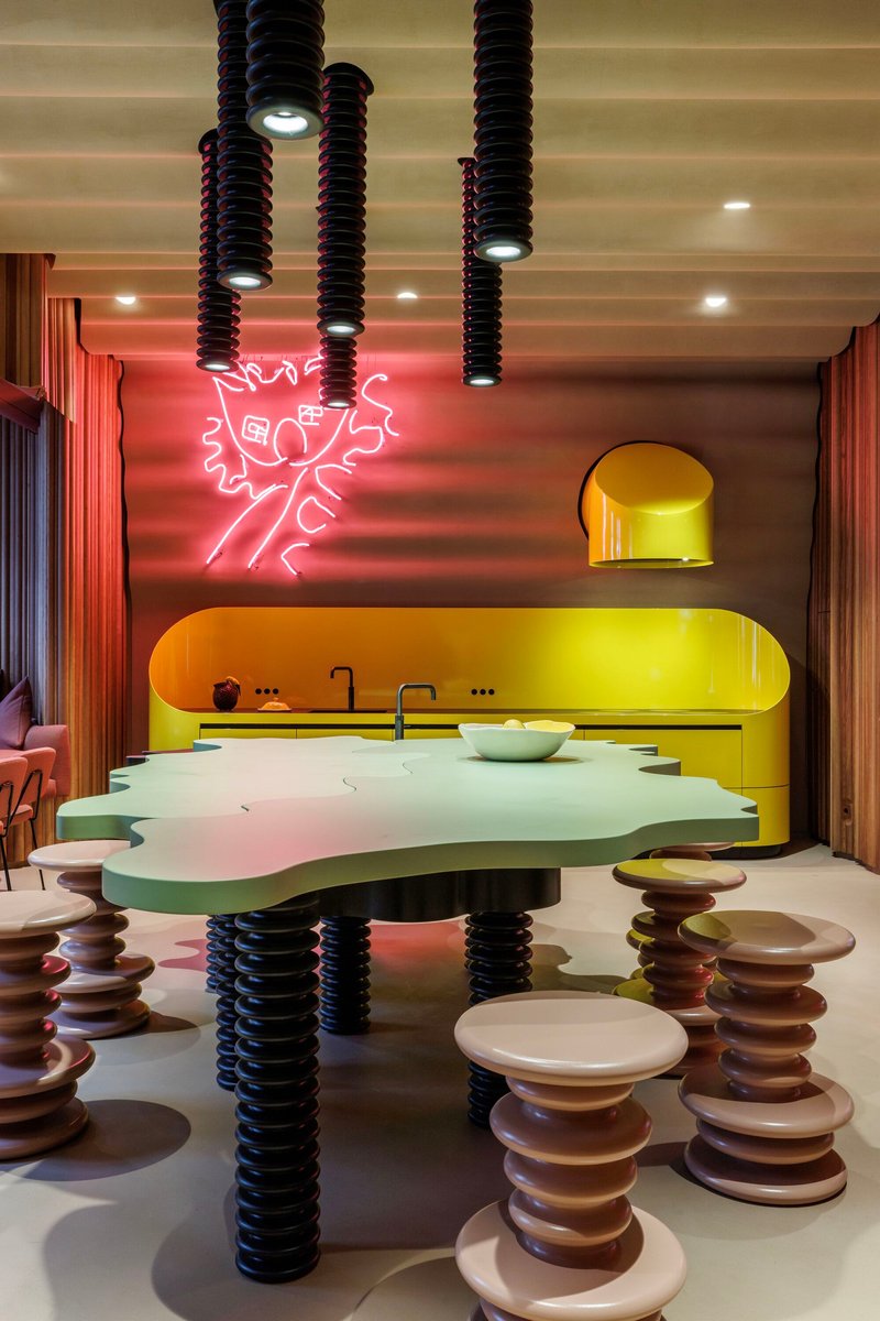 Open kitchen with yellow cabinetry, reflective floor, stacked disc stools, and pink neon wall art
