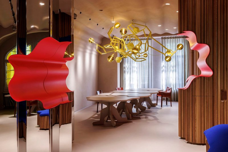 Interior view through red sculptural partition into dining area with gold geometric chandelier above timber table