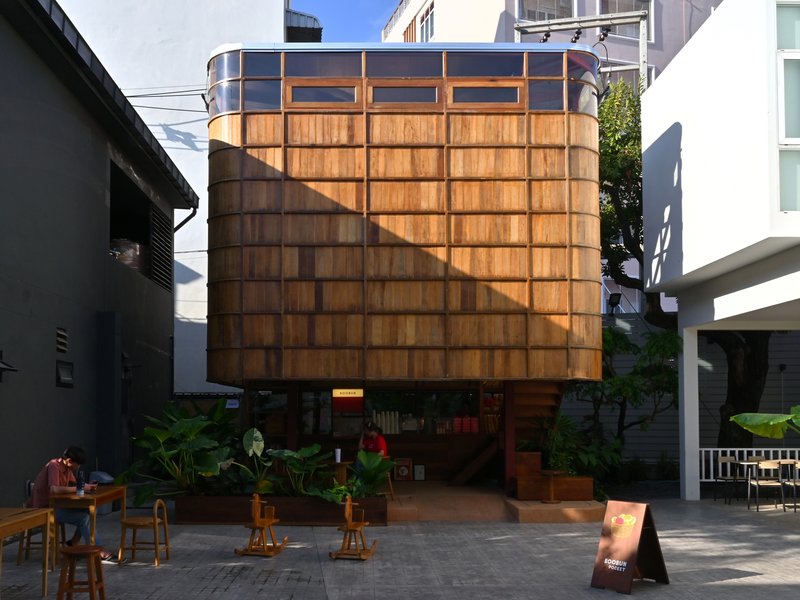 Elevated timber-clad volume on pilotis above a courtyard with potted plants and outdoor seating in afternoon light