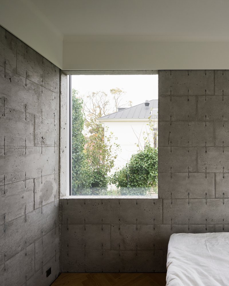 Bedroom corner with board-formed concrete walls and window overlooking trees and neighboring rooftop