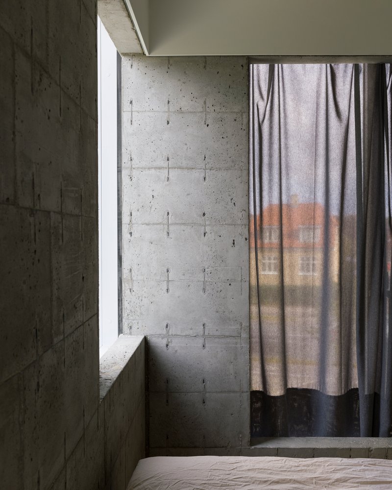Bedroom corner with board-formed concrete walls, tall window slot, and draped curtain filtering soft daylight