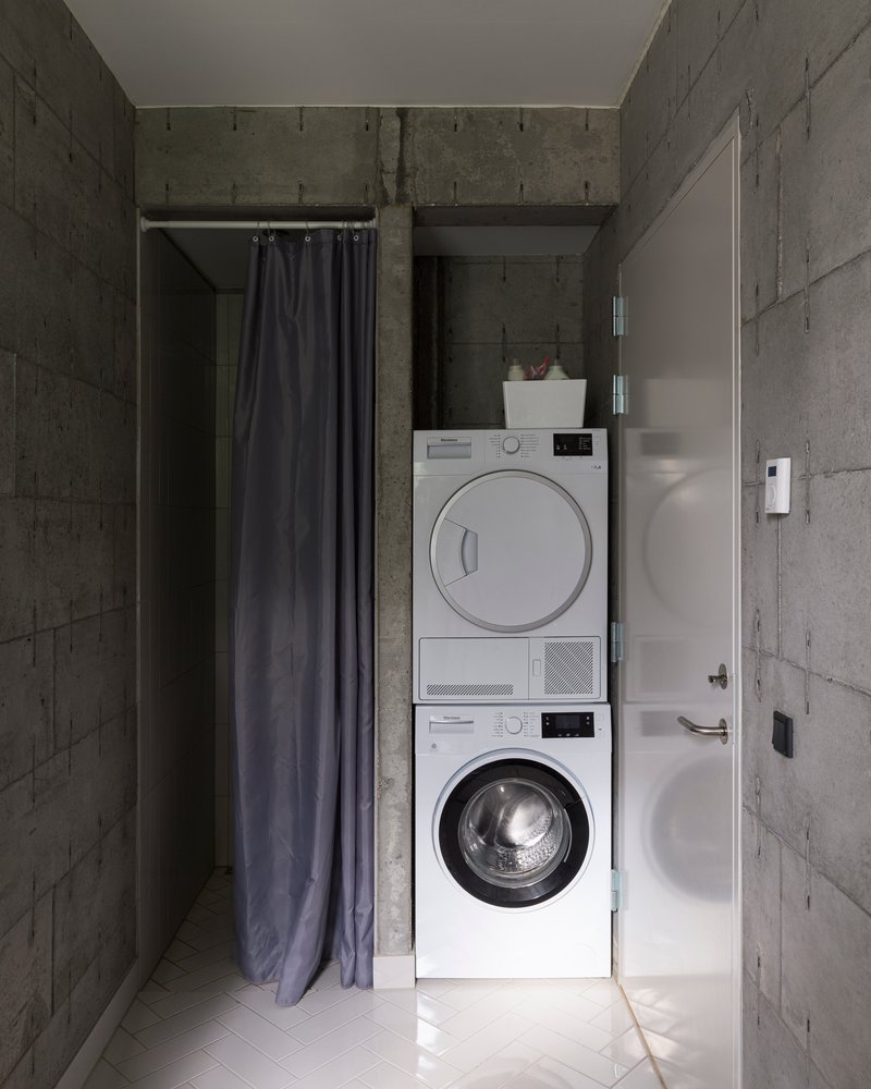 Compact laundry alcove with stacked washer and dryer flanked by dark curtains and concrete walls