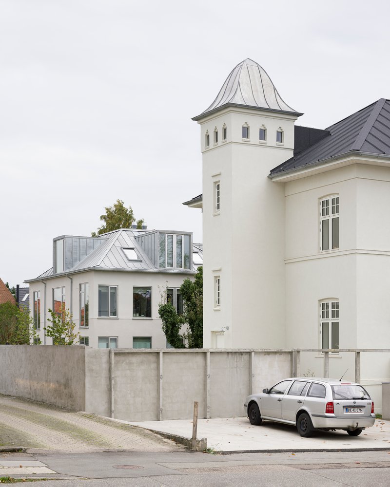 Street view of white tower with pointed roof beside zinc-clad addition behind concrete retaining wall